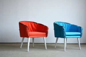 Dos sillones con cojines coloridos, uno naranja y otro azul, sobre suelo gris y pared blanca.